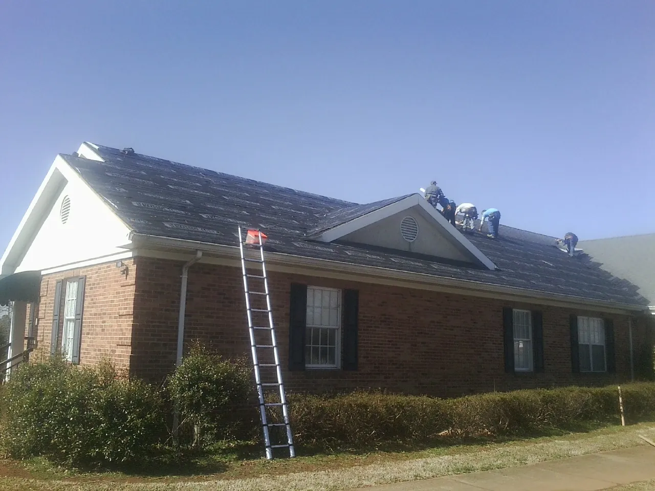 Crew tearing shingles off a brick ranch roof with a ladder set — Emergency Roof Repair work by our crew in Iona
