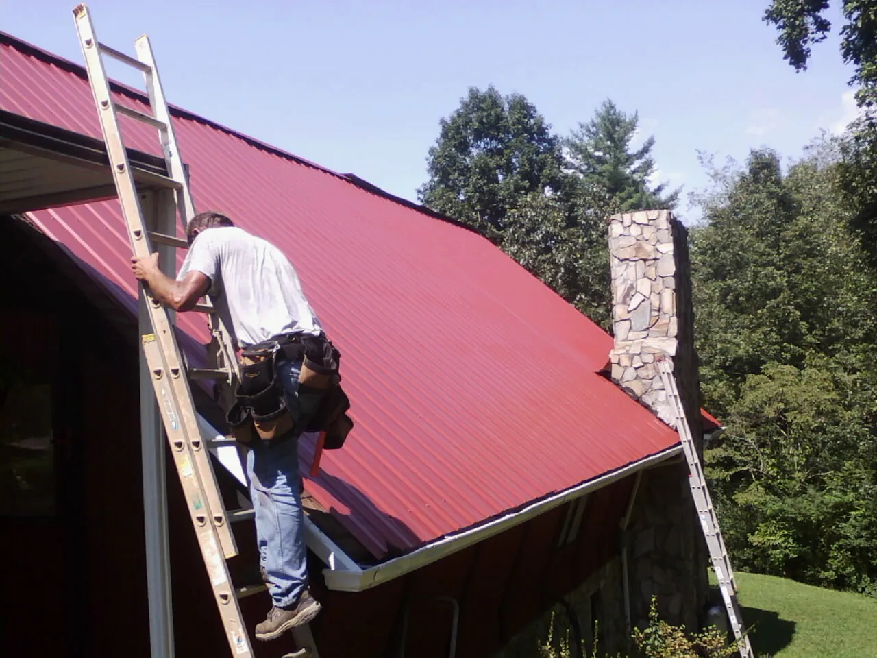 Roofer on a ladder beside a red metal roof and stone chimney — Soffit & Fascia Repair by Roofing Brothers in Iona